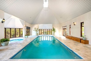 A large indoor swimming pool with a curved design, surrounded by a tiled floor and walls, with large windows providing natural light and views of greenery outside.