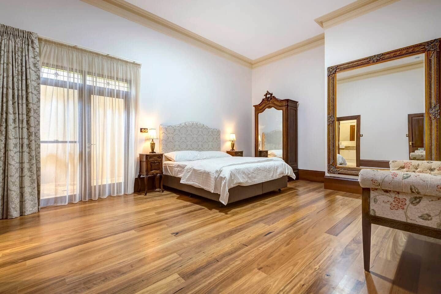 Une chambre d’hôtel avec un plancher en bois, un lit recouvert de draps blancs, une table de chevet avec une lampe, un grand miroir et un fauteuil à motifs floraux.