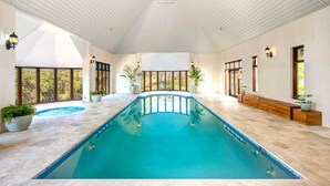 A large indoor swimming pool with a curved design, surrounded by a tiled floor and walls, with large windows providing natural light and views of greenery outside.