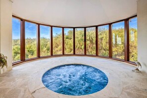 A hot tub in a room with large windows overlooking a forested area.