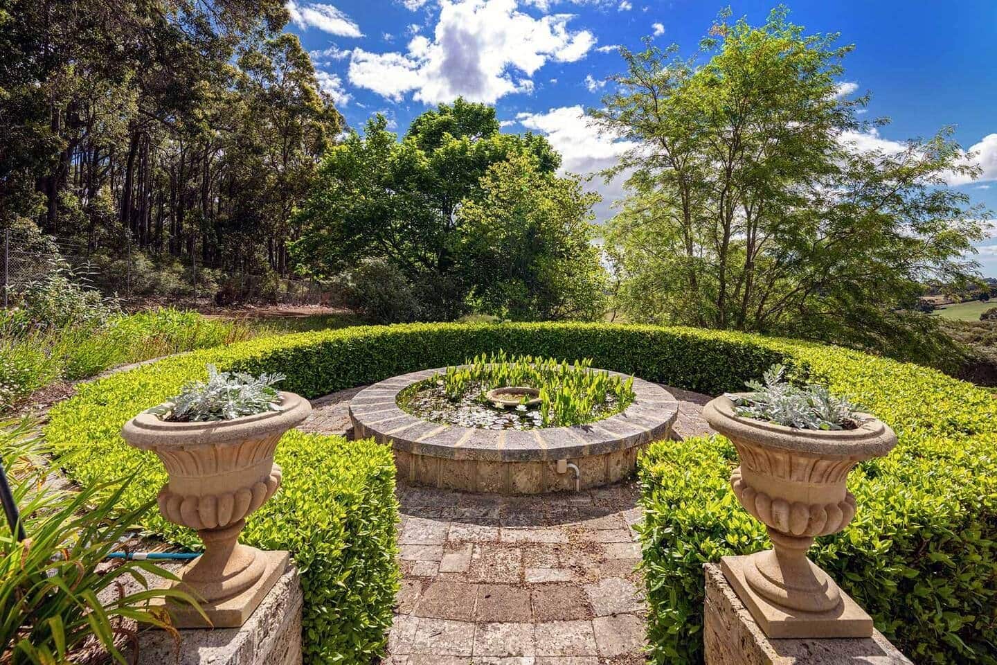 Un jardin aménagé avec une fontaine circulaire en pierre, bordé de haies soigneusement taillées et de plantes en pot.