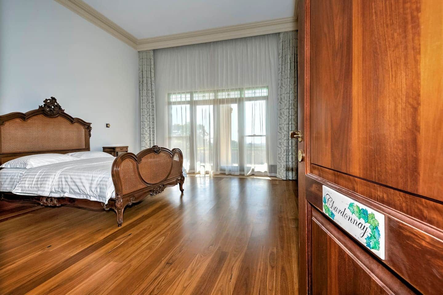 A spacious bedroom with a large bed, a wooden wardrobe, and a view through a window with sheer curtains.