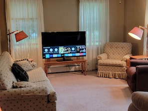 Una sala de estar con televisor de pantalla plana, dos sofás estampados, una mesa de centro de madera y una lámpara de pie.