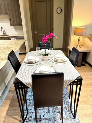 A dining area with a table set for four, chairs, a potted orchid, and a clock on the wall. - Stunning Pool View 2 Bedroom Apartment (Spring)
