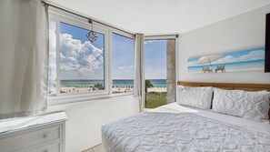 Un dormitorio con cama, mesita de noche, ventana con vista a la playa y un cuadro de una escena costera.