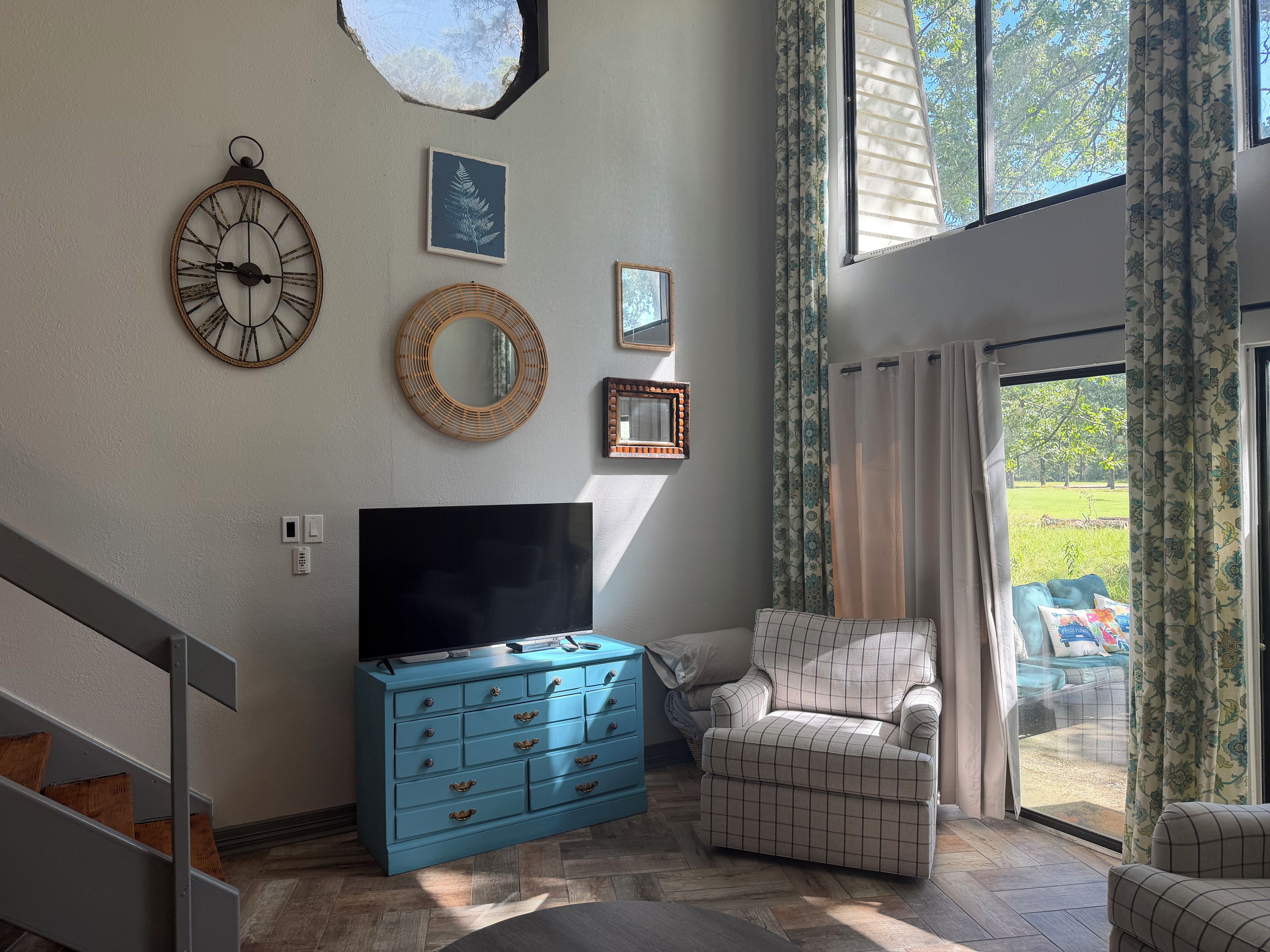 A living room with a flat-screen TV, a blue dresser, a patterned armchair, and a large window with curtains.
