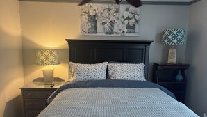 Un dormitorio con una cama, una mesita de noche, una lámpara, un ventilador de techo y una pared con un cuadro floral.
