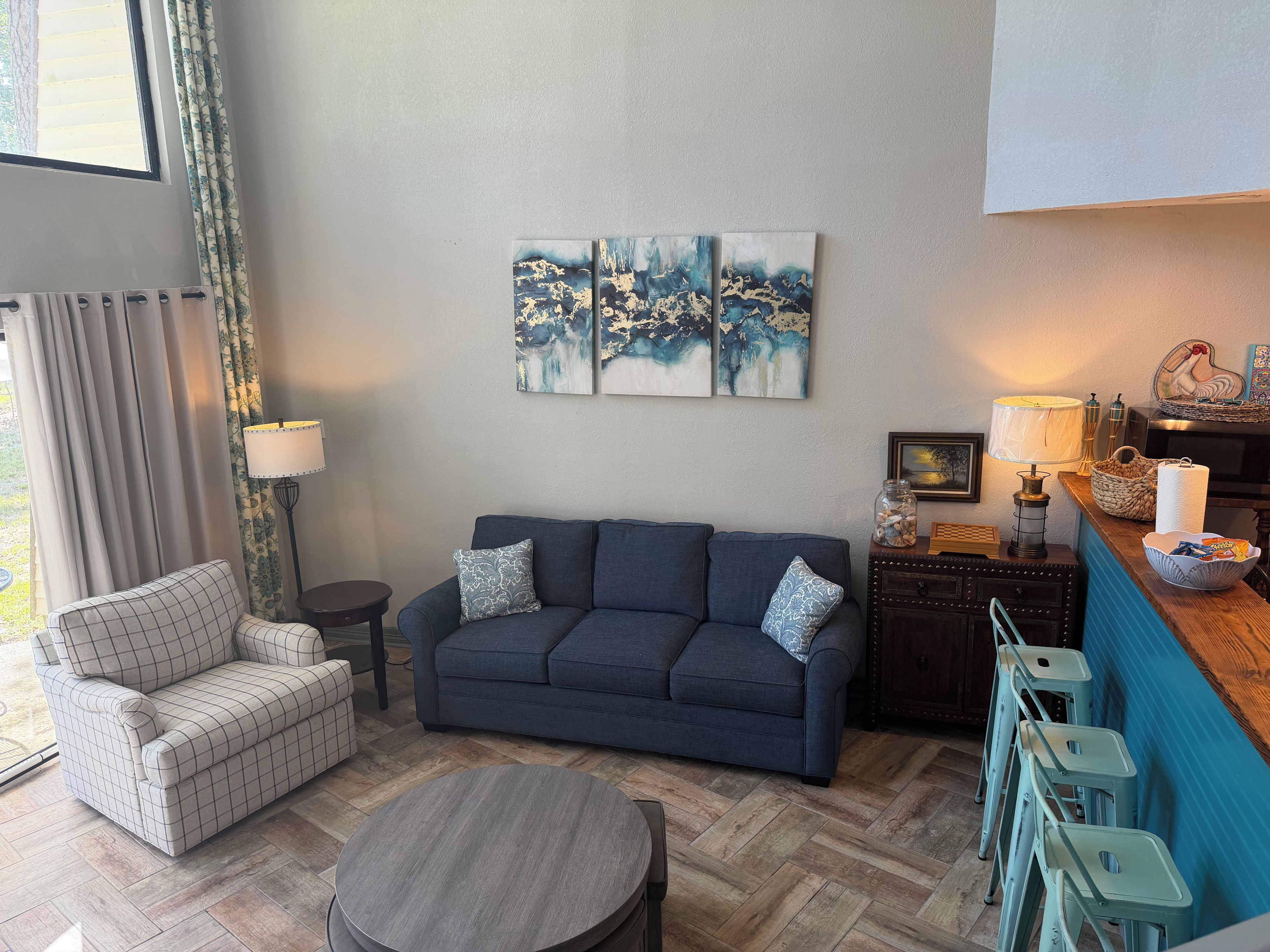 Een woonkamer met een bank, fauteuil en een rond salontafeltje. Twee schilderijen hangen aan de muur, er is een lamp en een klein tafeltje met een mand en een pot.