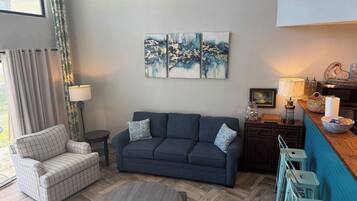 Een woonkamer met een bank, fauteuil en een rond salontafeltje. Twee schilderijen hangen aan de muur, er is een lamp en een klein tafeltje met een mand en een pot.