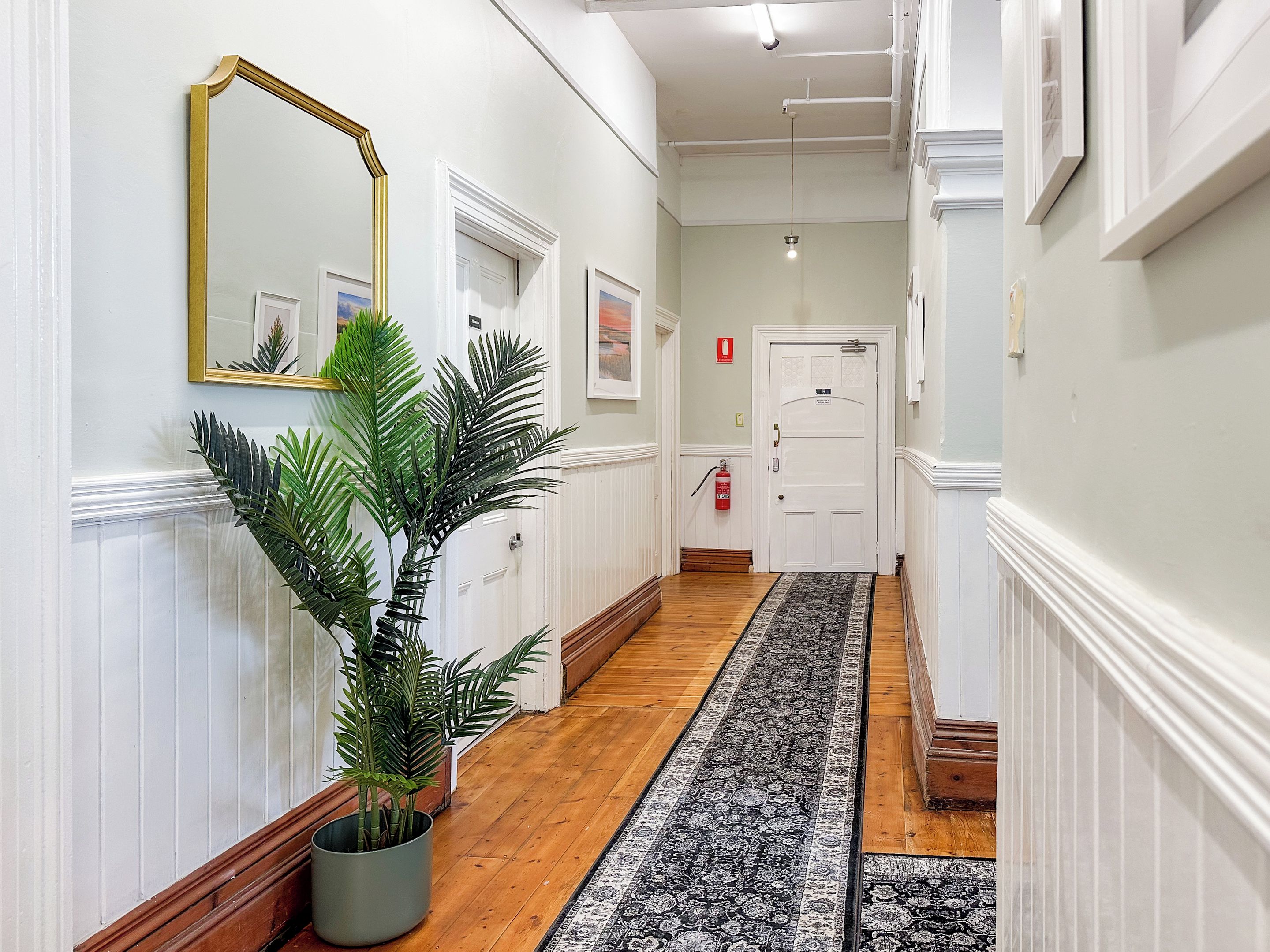 Un couloir doté d’un plancher en bois, d’un tapis à motifs noir et blanc, d’une plante en pot, d’un miroir avec un cadre doré et d’une porte blanche sur laquelle est fixé un extincteur.