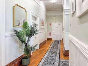 A hallway with a wooden floor, a black and white patterned rug, a potted plant, a mirror with a gold frame, and a white door with a fire extinguisher.