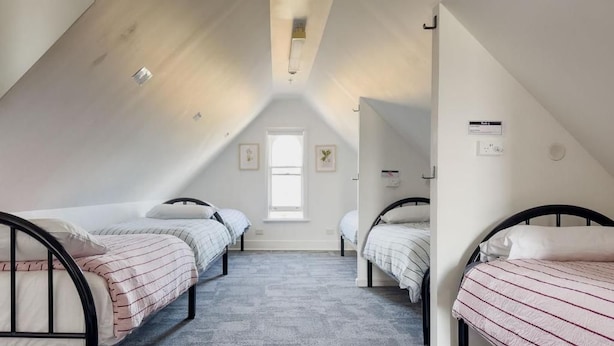 A room with three beds arranged in a U-shape, a window at the far end, and a ceiling with a light fixture. - The Fenway Hostel (Invermay)