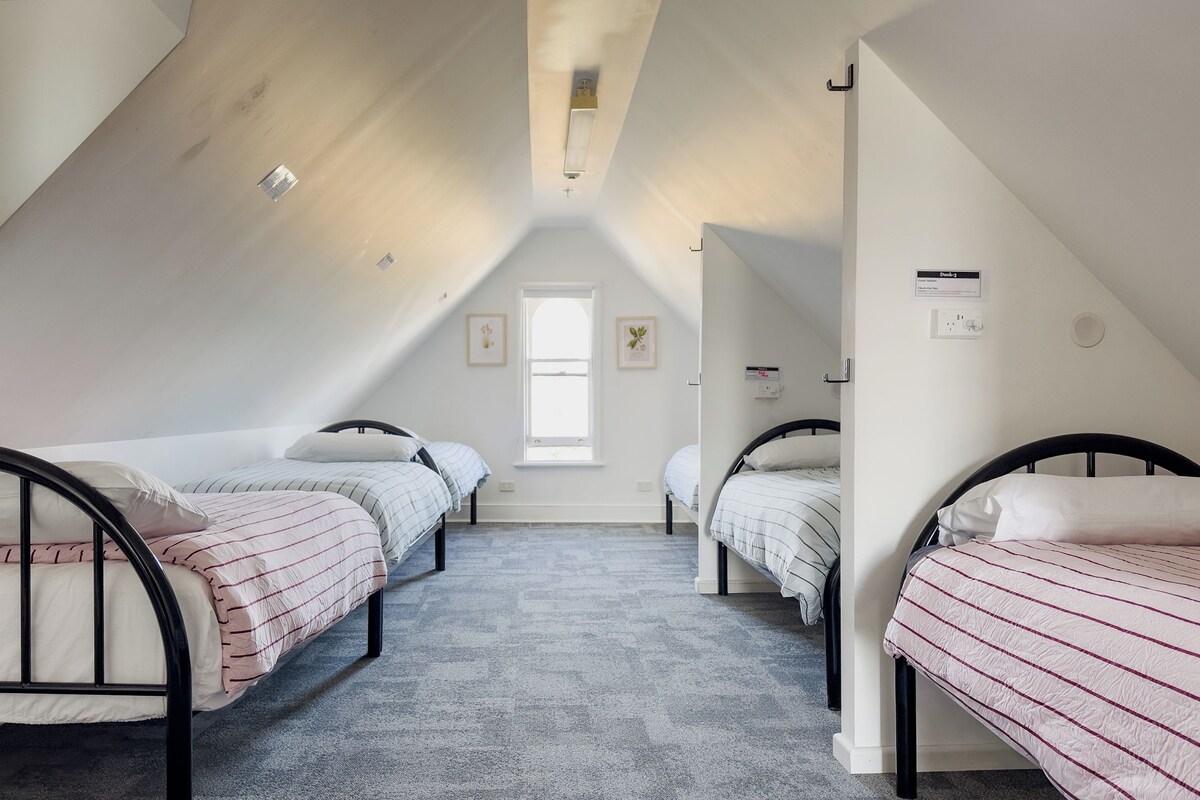 Quarto em dormitório com duas camas de solteiro, uma janela e um ventilador de teto.