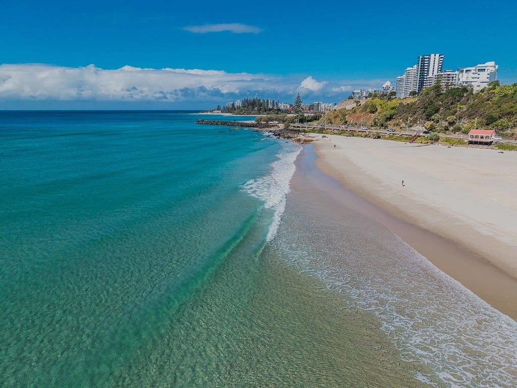 Uma praia com águas turquesa cristalinas, uma faixa de areia e edifícios ao fundo.