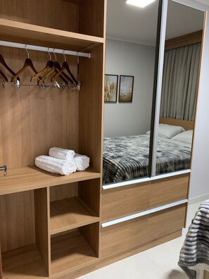 Een hotelkamer met een houten garderobe, een bed met een grijs gestreept dekbed en een grote spiegel die de kamer weerspiegelt.