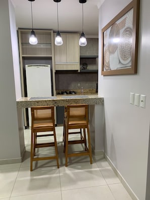 A kitchen with a central island, two bar stools, pendant lights, a refrigerator, and a wall-mounted decorative piece.