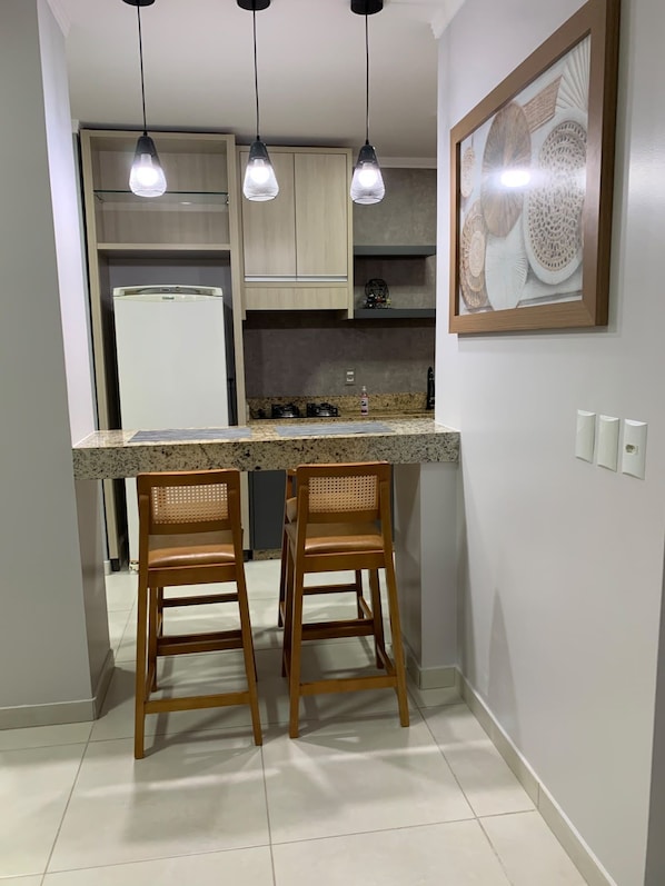 A kitchen with a central island, two bar stools, pendant lights, a refrigerator, and a wall-mounted decorative piece.