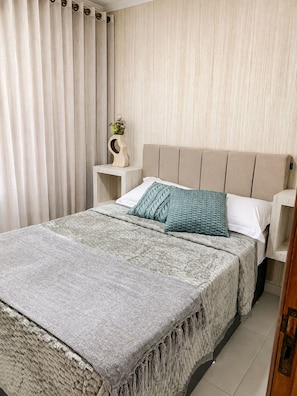Een slaapkamer met een bed, nachtkastje, een wandplank en een gordijn.