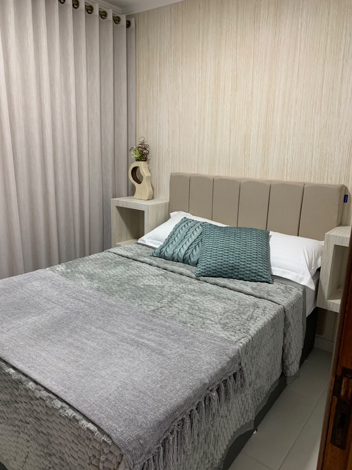 A neatly made bed with a grey blanket and pillows, a bedside table with a vase, and a curtain on the window.