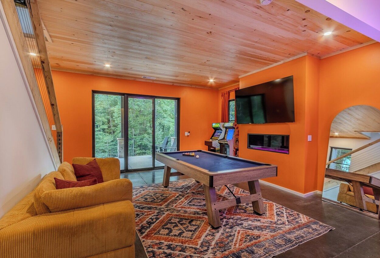 A room with a pool table, a sofa, a television mounted on the wall, and a staircase.