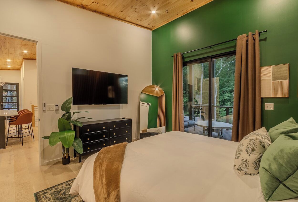 A bedroom with a large bed, a TV mounted on the wall, a dresser, and a window with curtains.