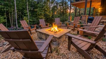 A circular fire pit surrounded by wooden Adirondack chairs, set on a gravel surface with a backdrop of trees and a wooden structure.