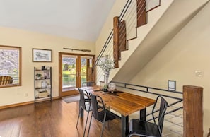 A dining area with a wooden table and chairs, a staircase with wooden railings, and a view of the outdoors through a glass door.