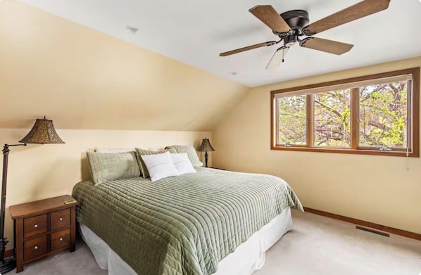 A bedroom with a bed, nightstands, a ceiling fan, and a window with a view of trees.