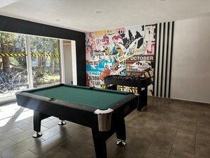 A pool table in a room with a large window, a wall with various posters, and a black foosball table. - Espaço Bosque Pucón: Nature Near the City Center (Pucon)
