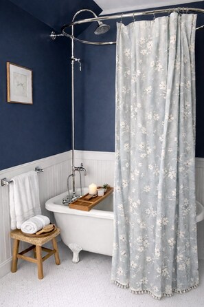 Une salle de bain avec une baignoire sur pieds blanche, un tabouret en bois, une serviette blanche et un rideau de douche à motifs floraux.