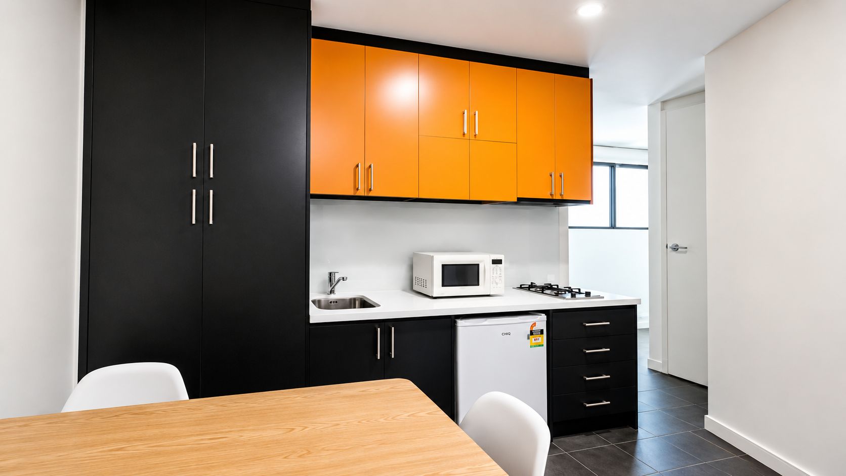 Une cuisine compacte avec des meubles noirs et orange, un plan de travail blanc, un four à micro-ondes, un petit réfrigérateur et une table à manger en bois avec des chaises.
