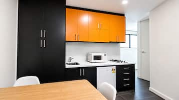 Cozinha compacta com armários preto e laranja, bancada branca, micro-ondas, pequena geladeira e mesa de jantar de madeira com cadeiras.