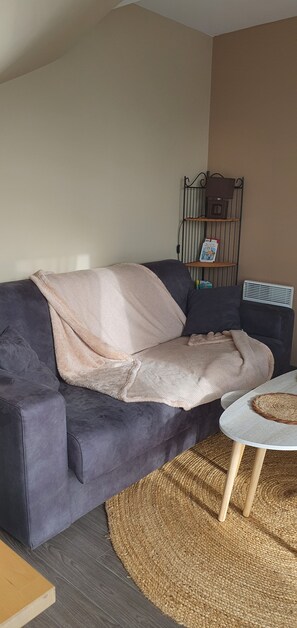 A living room with a dark-colored sofa, a light-colored throw blanket, a small round table with a woven placemat, and a wall-mounted shelf with books and a small decorative item. - Vrbo Property (Fort-Mahon-Plage)
