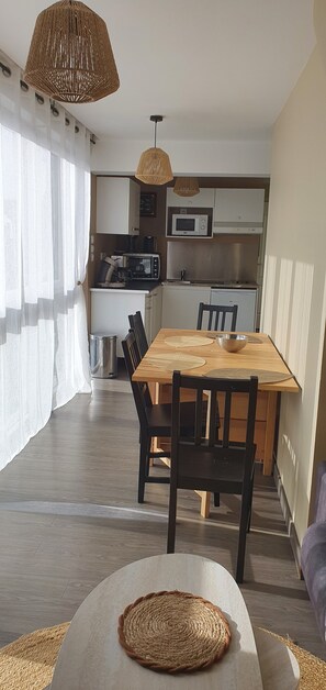 A modern kitchen with a dining area, featuring a wooden table and chairs, a woven pendant light, and a view of the living area. - Vrbo Property (Fort-Mahon-Plage)