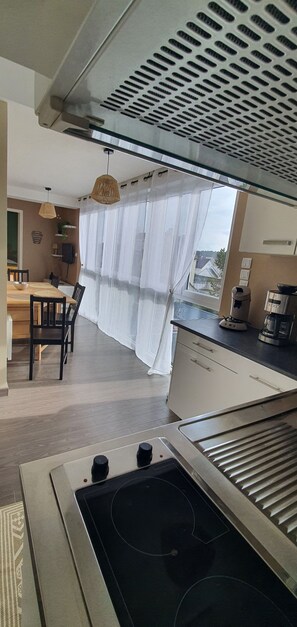 A modern kitchen with a built-in stove, a dining area with a table and chairs, and a view through sheer curtains. - Vrbo Property (Fort-Mahon-Plage)