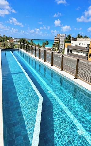 Ein langer, rechteckiger Swimmingpool mit klarem, blauem Wasser, umgeben von einem Metallgeländer. Im Hintergrund sind Gebäude und ein strahlend blauer Himmel zu sehen.