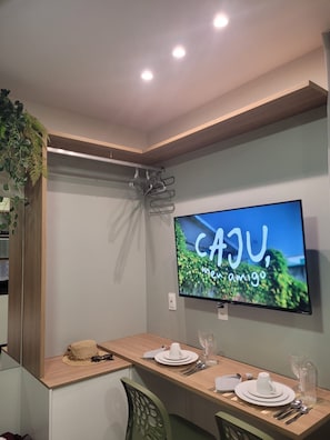 Ein moderner Essbereich mit einem Flachbildfernseher, der 'CAJU, amigo' an der Wand zeigt, einem Holztisch, der für zwei Personen gedeckt ist, und einer hängenden Pflanze.