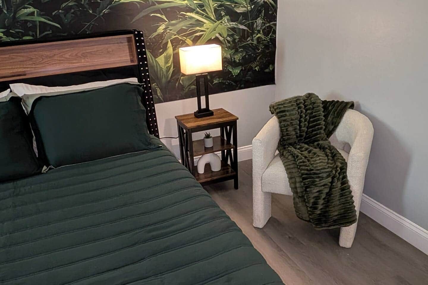 Ein Schlafzimmer mit einem Bett, einem Nachttisch mit Lampe, einem Sessel und einer Wand mit Pflanzenwandbild.