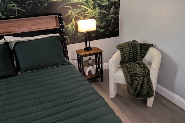 A bedroom with a bed, a nightstand with a lamp, a chair, and a wall with a plant mural.