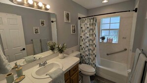 Ein Badezimmer mit Waschbecken, Toilette und Badewanne. Ein Fenster bietet einen Ausblick, eine Duschvorhang ist angebracht, und über dem Spiegel befinden sich Lampen.