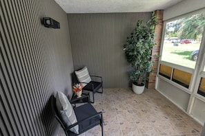 A small, enclosed patio area with striped walls, two chairs, a potted plant, and a view through a glass door.