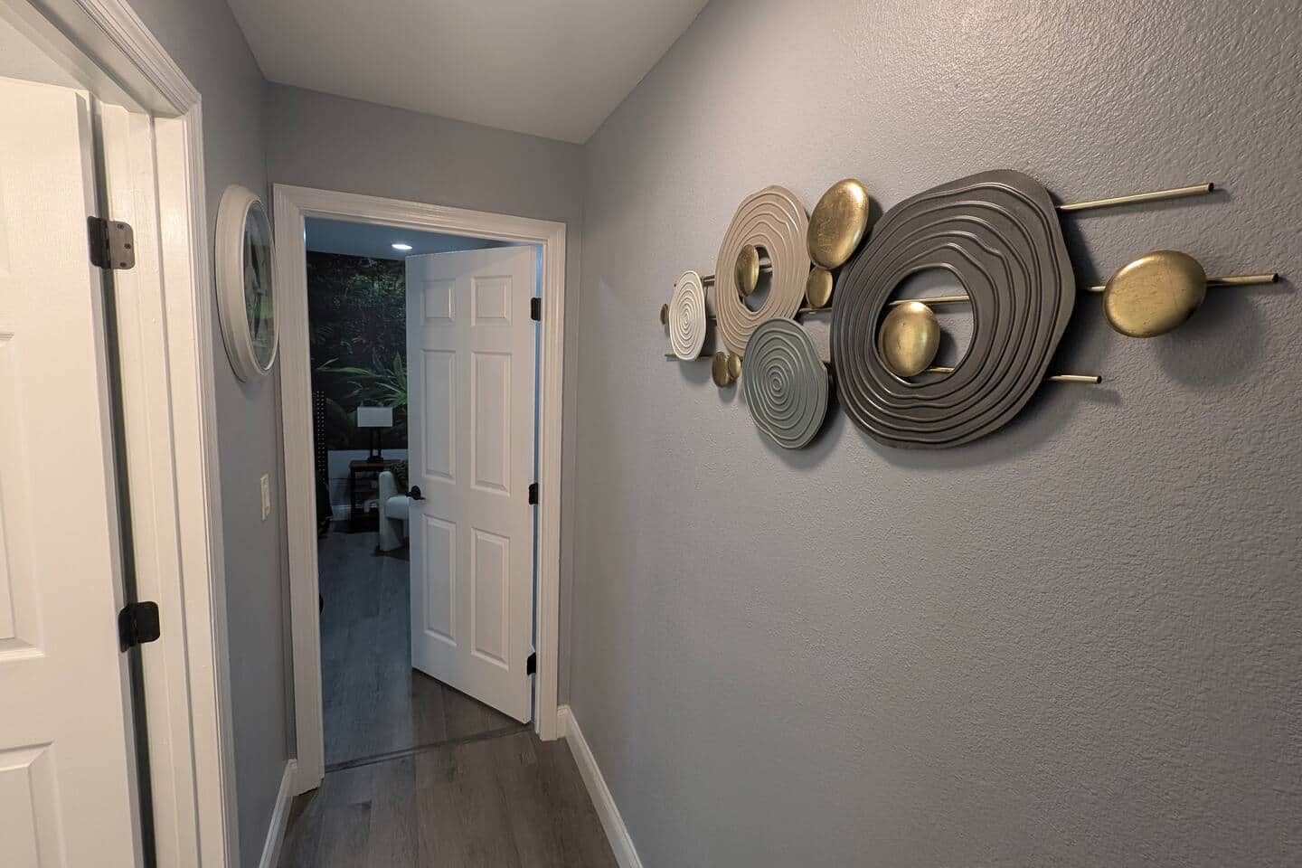 Ein Flur mit einer zurueckfuehrenden Tür zu einem Zimmer, einem an der Wand befestigten dekorativen Metallkunstwerk und einer Tür mit Griff.
