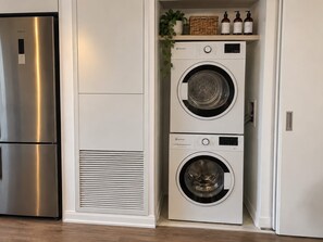 Cozinha compacta com geladeira de aço inoxidável, máquina de lavar e secadora embutidas e uma prateleira com produtos de higiene pessoal.