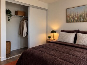 A bedroom with a bed, a nightstand with a lamp, a painting, a closet with clothes and baskets, and a potted plant. - Modern Downtown Toronto Studio with Balcony & Stunning City Views. (Toronto)