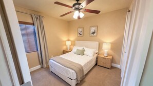 Une chambre à coucher avec un lit, deux tables de chevet avec des lampes, un ventilateur de plafond et une fenêtre avec des rideaux.