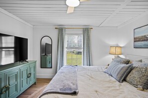 Ein Schlafzimmer mit einem Bett, einem Fernseher, einem Spiegel, einem Fenster mit Vorhängen, einer Lampe und einem Bild an der Wand.