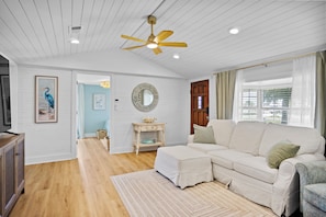 A spacious living room with a white sofa, a ceiling fan, and a large window with curtains. - 3BR Coastal Cottage - Steps to St. Joseph Bay (Port St. Joe)