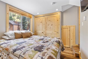 A bedroom with a large bed, a window with blinds, a wooden wardrobe, and a small wooden chair.