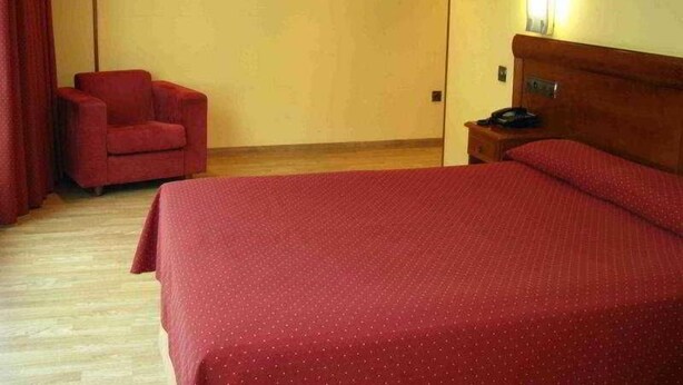 Quarto de hotel com cama, cadeira, mesa de cabeceira e luminária na parede.