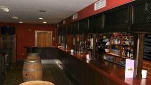 Uma área de bar com barris de madeira, um balcão com garrafas e uma parede vermelha.